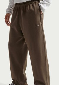 Pantalon de survêtement marron avec une ceinture élastique, des poches latérales et un petit logo Nike blanc sur la jambe gauche. Tissu en coton lisse et coupe droite.