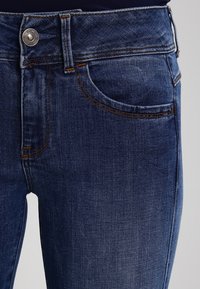 Jeans de mezclilla azul oscuro con una textura suave, diseño de cinco bolsillos, acentos de costura visibles y un cierre de botón plateado en la cintura.