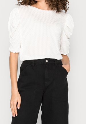 Witte textuurblouse met opgeblazen mouwen, gecombineerd met hooggetailleerde zwarte broek met steekzakken aan de voorkant en een knoopsluiting.