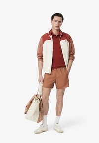 Idrottsklädsel som består av en vit och rostfärgad jacka, röd pikétröja, bruna shorts och vita sneakers. Tillbehören inkluderar en mönstrad tote-väska.