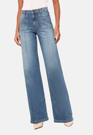 Femeie purtând blugi albaștri cu talie înaltă și picior larg, cu buzunare mari în față și pantofi bej cu vârf ascuțit, stând în fața unui fundal simplu.