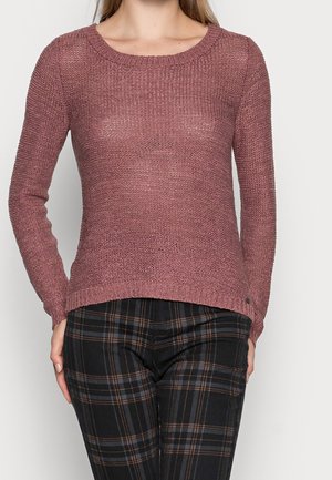 Sweter w kolorze przykurzonego różu, dzianinowa struktura z prążkowanym dekoltem i mankietami. W połączeniu z czarnymi spodniami w kratę z brązowymi akcentami i dopasowanym krojem.