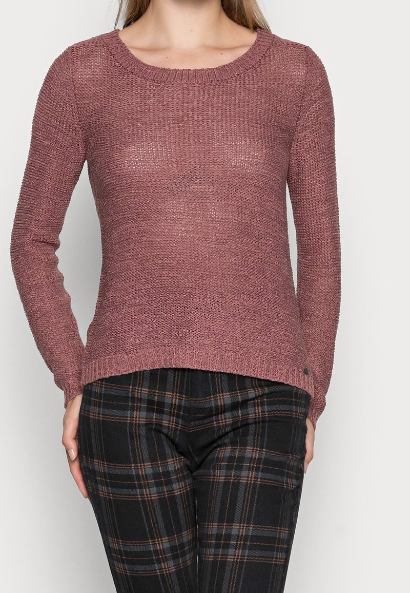 Pullover in staubrosa, gestrickt mit Rippkragen und -bündchen. Kombiniert mit schwarzen Karohosen mit braunen Akzenten und schmalem Schnitt.