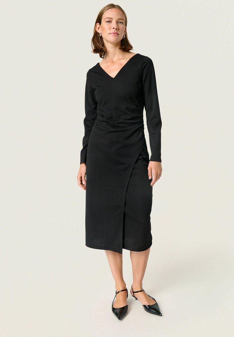 Vestido negro de manga larga con escote en V, cinturilla ajustada y largo hasta la rodilla. Presenta un diseño cruzado y una textura de tela suave. Combinado con zapatos de tacón negros.