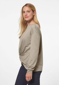 Hellbeige Langarmoberteil mit lockerer Passform und einem Knoten-Detail in der Taille. Der Stoff wirkt glatt und weich. Kombiniert mit dunklen Hosen.