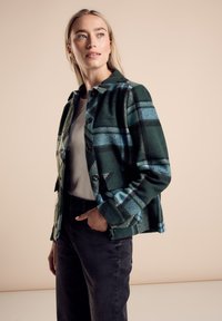 Groen en blauw geruite wollen jas met een kraag, frontknopen en twee zijzakken. Gecombineerd met een lichte top en zwarte jeans.