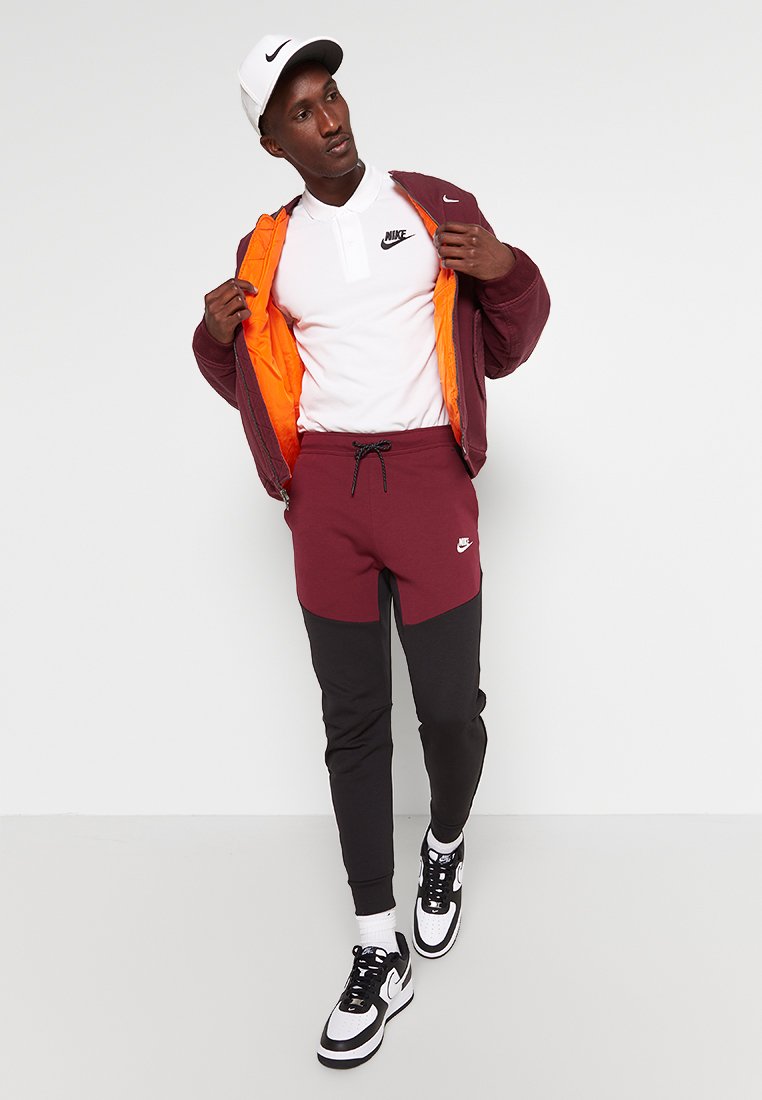 Giacca Nike di color bordeaux con fodera arancione, polo bianca e pantaloni jogger bordeaux e neri. Sneakers bianche e nere completano l'outfit.