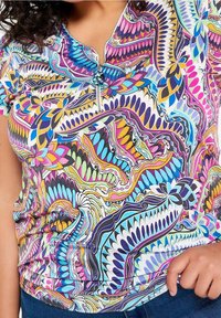 Donna che indossa un top colorato con un motivo astratto, con una cerniera corta all'collo e jeans blu, con la mano appoggiata vicino alla tasca.