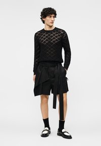 Άνδρας που φοράει μαύρο πουλόβερ με ανάγλυφα μοτίβα, ασύμμετρα μαύρα σορτς με κρεμασμένο ύφασμα, μαύρες κάλτσες και ασπρόμαυρα μοκασίνια, όρθιος.