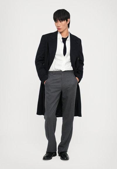 Cappotto nero, camicia bianca, pantaloni grigi e scarpe nere. Il cappotto presenta un design lungo; i pantaloni hanno una texture liscia.
