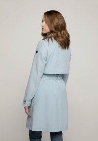 Lichtblauwe trenchcoat met een ceintuur in de taille, lange mouwen en knoopaccenten. Heeft een cropped ontwerp aan de achterkant en zijzakken.