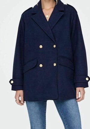 Femme portant un manteau bleu marine croisé avec des boutons dorés, des poches à rabat et un jean sur un fond uni.