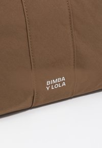 Bolsa de tela marrón con costuras verticales y el logotipo blanco "BIMBA Y LOLA" en la parte inferior derecha. Presenta una textura suave y un diseño sencillo.