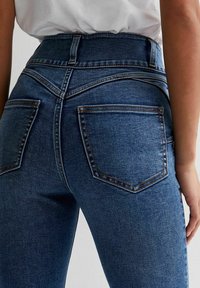 Jeans de mezclilla azul de cintura alta con una forma ajustada, que cuentan con dos bolsillos traseros y detalles de costura visibles. Textura de tela suave.