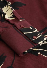 Poignet de chemise manches longues couleur marron avec imprimé floral abstrait noir et crème et deux boutons marron sur la manche.