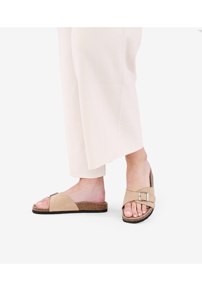 Pieds portant des sandales beige à bout ouvert avec semelles en liège et brides à boucle, associés à un pantalon large couleur crème clair.