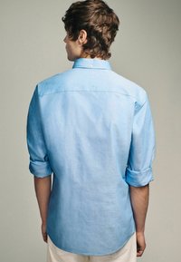 Helles blaues Button-up-Hemd aus Baumwolle, mit hochgerollten Ärmeln, abgerundetem Saum, schlichtem Design und sichtbaren Nähten, getragen mit beigen Hosen.
