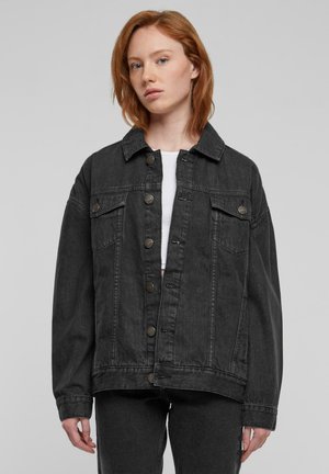 Giovane con capelli rossi che indossa una giacca di jeans nera oversize sopra una maglietta bianca su sfondo semplice.