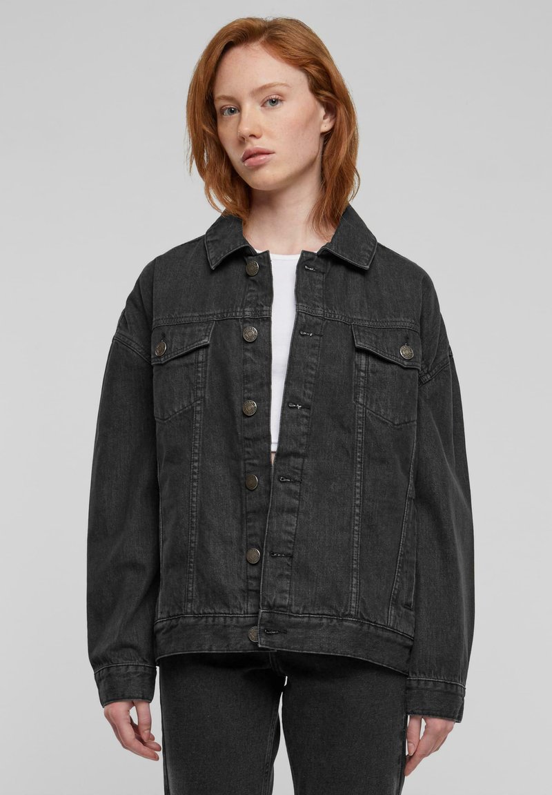 Ung person med rødt hår iført oversized sort denimjakke over hvid top mod ensfarvet baggrund.