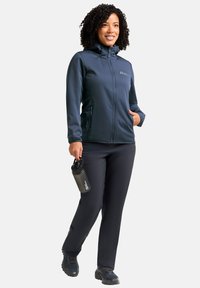 Vrouw loopt in een donkerblauwe softshell jas en broek, houdt een zwarte waterfles vast, glimlacht, op een witte achtergrond.