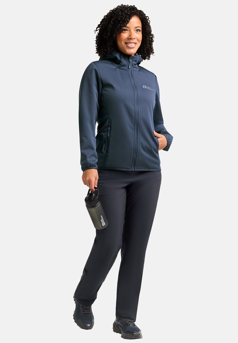 Vrouw loopt in een donkerblauwe softshell jas en broek, houdt een zwarte waterfles vast, glimlacht, op een witte achtergrond.