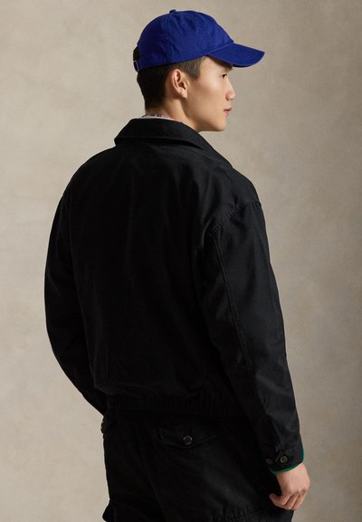 Sort jakke med afslappet pasform, der har krave, elastisk talje og lommer. Kombineret med en blå kasket mod en neutral baggrund.