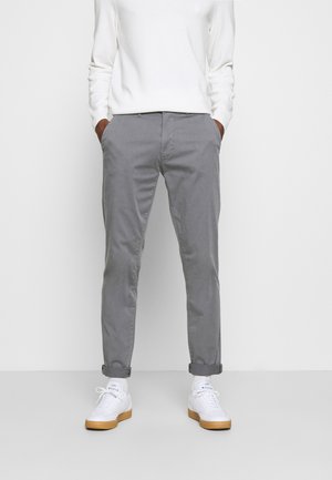 Persoon met grijze opgerolde broek, wit shirt met lange mouwen, witte sokken en witte sneakers met bruine zolen, staand met handen in de zakken.