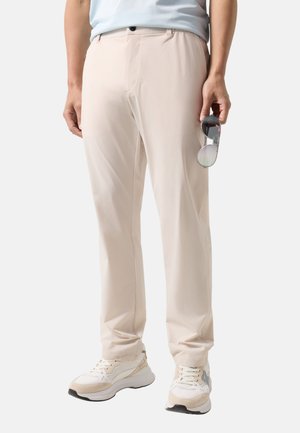 Mand iført beige bukser, hvide sneakers, lyseblå skjorte, der holder reflekterende solbriller i højre hånd mod ensfarvet baggrund.