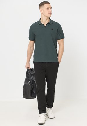 Uomo che indossa una polo verde scuro e pantaloni neri, che cammina tenendo una borsa da viaggio in pelle nera nella mano sinistra.