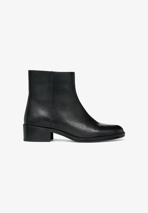 Stivale alla caviglia in pelle nera con punta arrotondata, tacco a blocco basso e dettagli di cucitura discreti lungo la suola. Texture liscia, design minimalista.