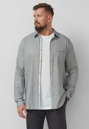 Mann mit Bart, der ein hellgraues Hemd über einem weißen T-Shirt und dunklen Hosen trägt, schaut nach links, eine Hand in der Tasche.