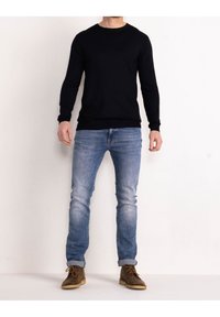 Uomo in piedi, che indossa un maglione nero a maniche lunghe, jeans chiari arrotolati e stivali marroni con lacci, su uno sfondo bianco semplice.