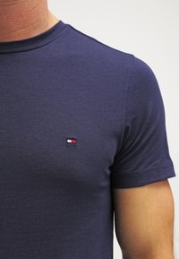 Primo piano di una persona che indossa una maglietta a maniche corte blu navy con un piccolo logo rettangolare rosso, bianco e nero sul petto.