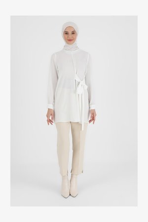 Tunique blanche à manches longues avec une ceinture à la taille, associée à un pantalon beige ajusté et des bottines à talons pointus. Tissu lisse avec un design léger.