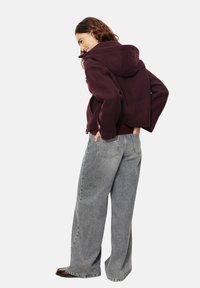 Veste à capuche bordeaux au cut décontracté et avec des manches larges, associée à un jean gris clair à coupe ample. La tenue présente des détails de couture uniques.