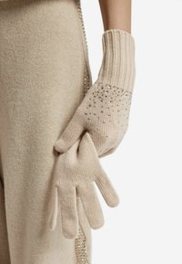 Beżowe dziergane rękawiczki z prążkowanymi mankietami i zdobieniami z małymi srebrzystymi akcentami po stronie dłoni. Miękka tekstura.
