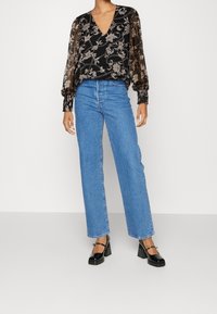 Blusa nera con motivo floreale, maniche lunghe trasparenti, abbinata a jeans a vita alta blu con gamba larga e scarpe con tacco nere.
