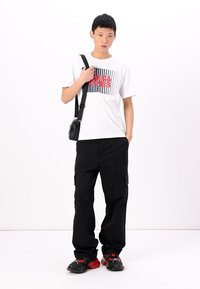 Weißes T-Shirt mit rotem und schwarzem grafischen Design, schwarze Cargo-Hosen und schwarze Schuhe mit roten Akzenten. Modell trägt eine kleine schwarze Tasche.