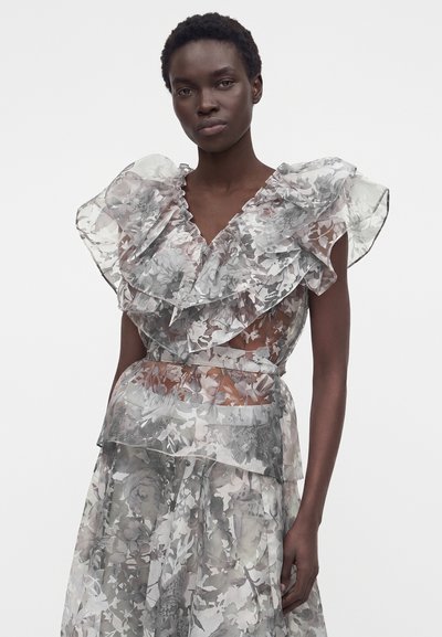 Γυναίκα που φορά διάφανο, με βολάν, λουλουδάτο φόρεμα με μεγάλο στρωματοποιημένο γιακά και ζώνη στη μέση, στέκεται μπροστά σε απλό ανοιχτό φόντο.