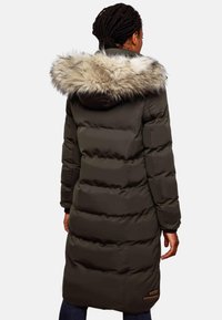 Long olive green puffer coat with a faux fur trim on the hood, quilted design, and side pockets, featuring a logo patch on the lower hem.