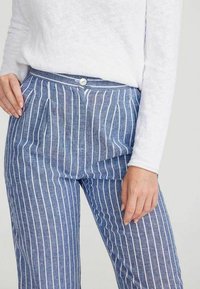 Blauwe gestreepte broek van zacht materiaal, met een knoopsluiting en plooien. Witte verticale strepen contrasteren met de blauwe stof.