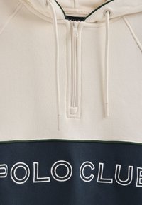 Off-white zip-up hoodie met een donkerblauw onderste paneel. Voorzien van geborduurde tekst "POLO CLUB". Zachte katoenmix stof met een gladde textuur.