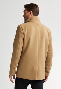 Cappotto beige in misto lana con colletto alto e design slanciato. Presenta una cucitura centrale sul retro e una vestibilità sartoriale.