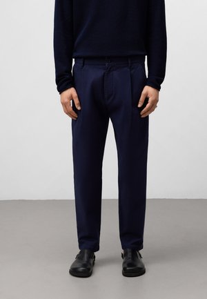 Navy blue trousers with a smooth texture, flat front, and pleated design. Worn with a dark long-sleeve top and black shoes.