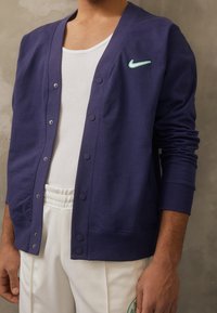Cardigan blu navy in cotone, con scollo a V, bottoni a pressione e piccolo logo Nike verde. Abbinato a una canottiera bianca e pantaloni.