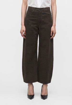 Femme portant un pantalon large taille haute marron foncé avec un imprimé léopard subtil et des talons noirs à bout pointu, debout devant un fond uni.