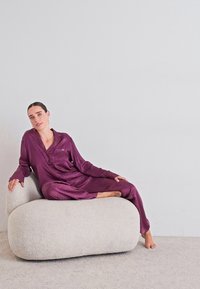 Moteris violetiniu satino pižamu atsilošusi ant apvalaus, tekstūruoto balto fotelio prie paprasto šviesiai pilko sienos.
