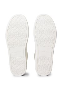 Balti sporta apavu zoles ar rakstainu rakstu un "Calvin Klein" logotipu iegravētu katras zoles centrā, redzamas no apakšas.