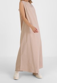 Lang, ærmeløs beige kjole med høj hals og sideslidser, parret med lyse sneakers. Glat, draperende stoftekstur.
