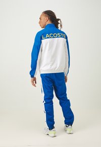 Zils un balts Lacoste sporta uzvalks, kas sastāv no jaku ar rāvējslēdzēju un biksēm ar kontrastējošām svītrām un lielu dzeltenu logo aizmugurē.